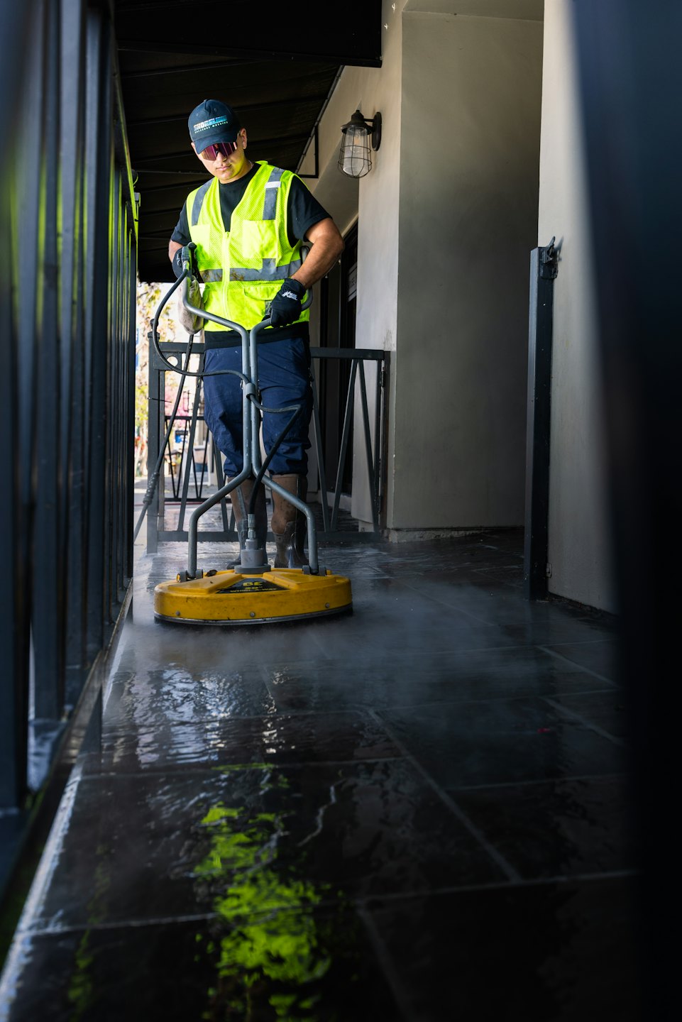Man in high-visibility vest power washes outdoor tiled floor.